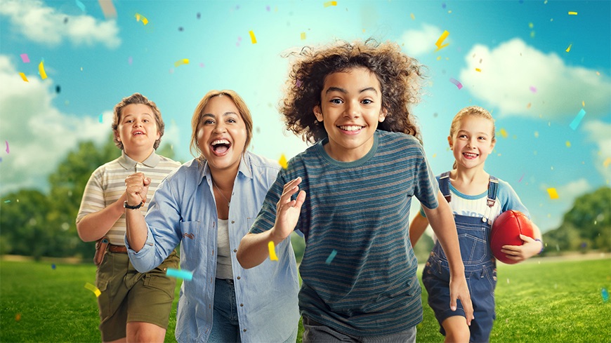 A group of four children and one adult joyfully run towards the camera, with smiles and laughter. Confetti falls around them, and they are set against a bright blue sky and green grass, creating a lively, celebratory atmosphere.