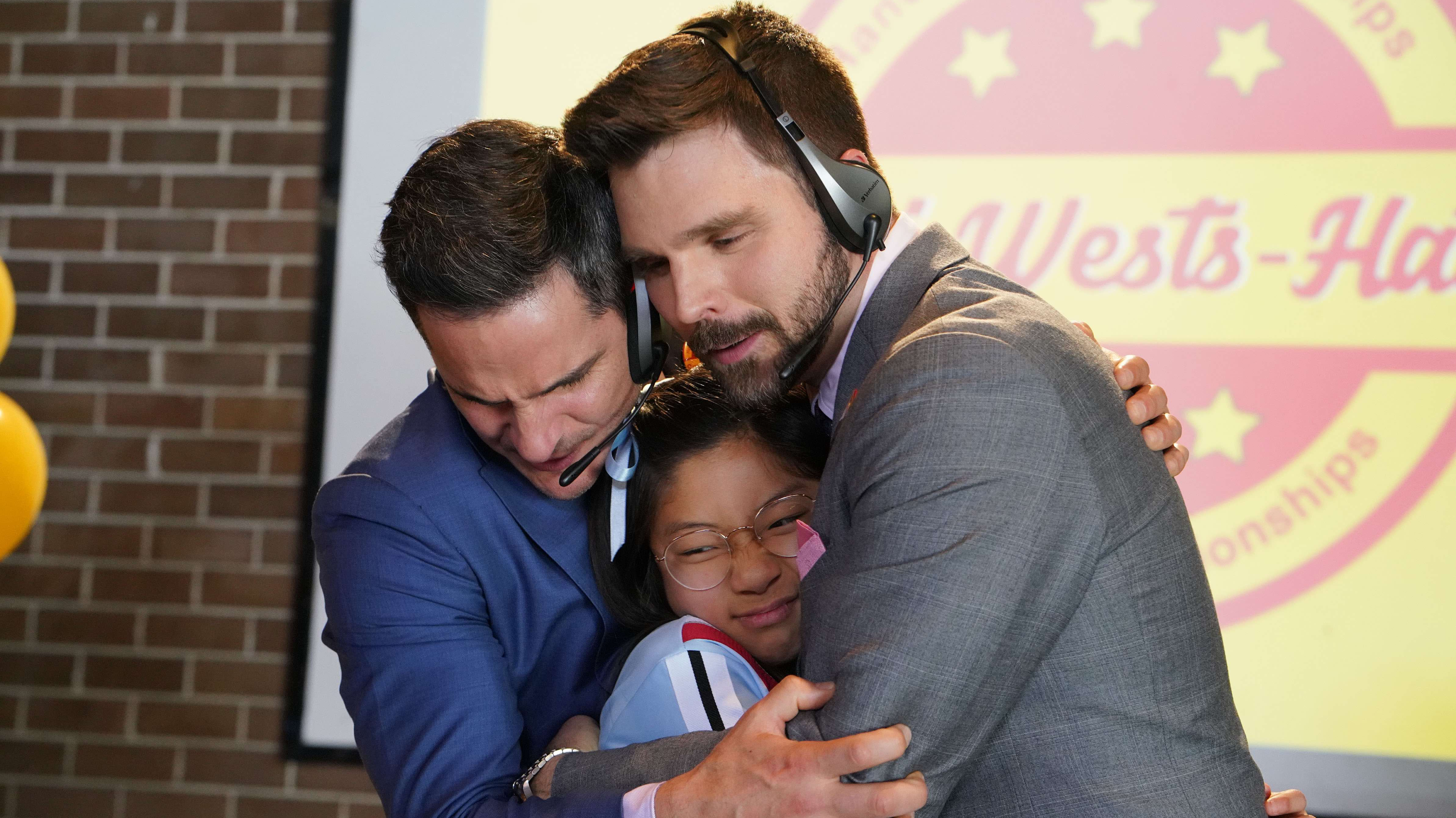 Hardball Two men wearing headsets embrace a young girl with glasses in a warm, supportive group hug. The background features a colorful logo for a championship event, suggesting a moment of celebration or achievement.