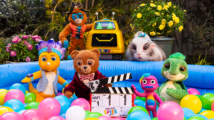 Knee_High_Spies_s2_ _Toys_(Photo_Sarah_Enticknap)_870x489px A group of colorful plush characters gathers around a blue inflatable pool filled with pastel-colored balls. Among them are a bear in a red jacket, a yellow creature with a blue mohawk, and a green crocodile, all posing cheerfully with a film clapperboard. In the background, a robot and a bunny-like creature can be seen, along with bright yellow flowers.